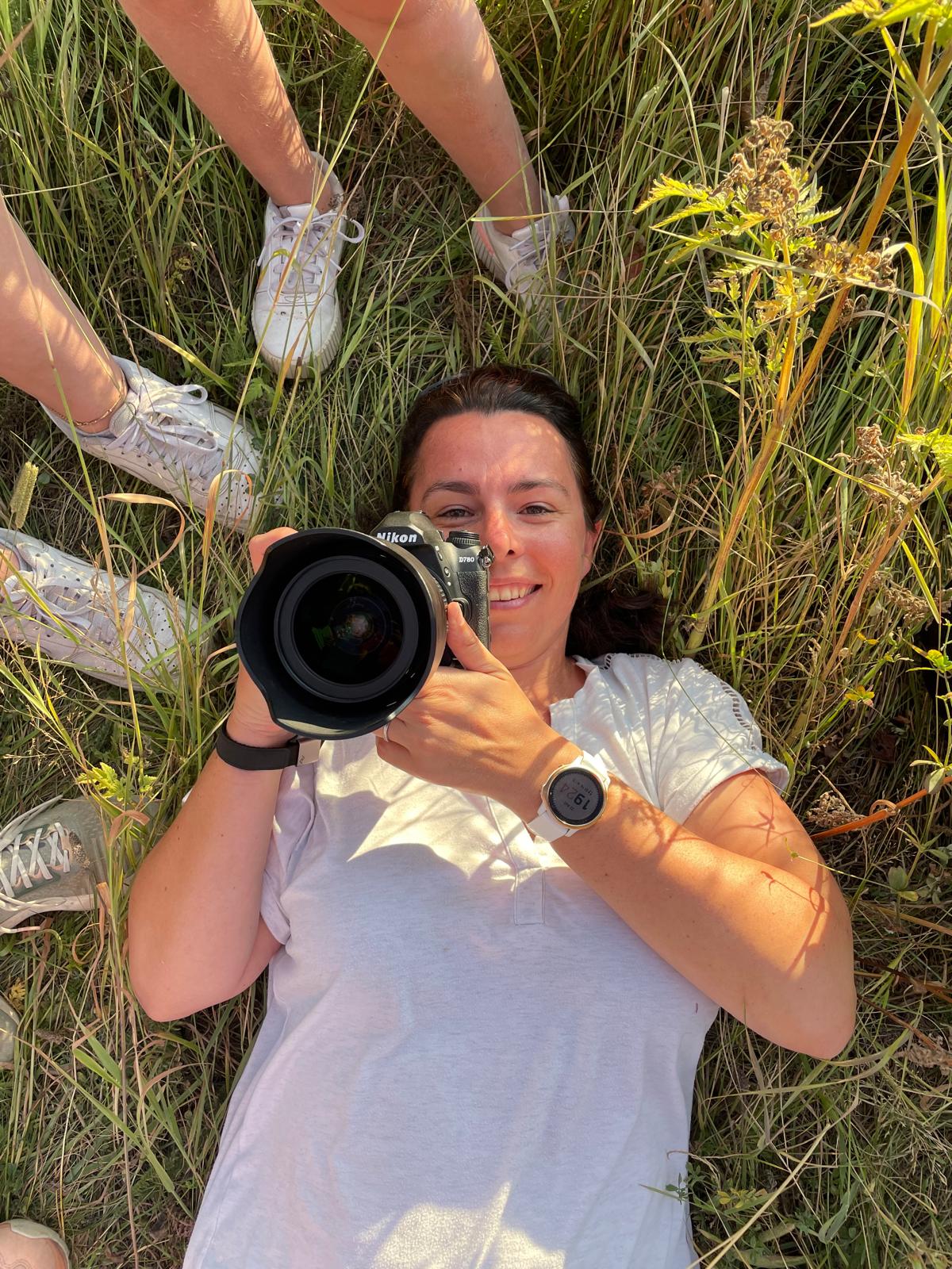 Portrait d'Aurélie Follain allongée dans l'herbe, appareil photo en main, lors d'un shooting en extérieur