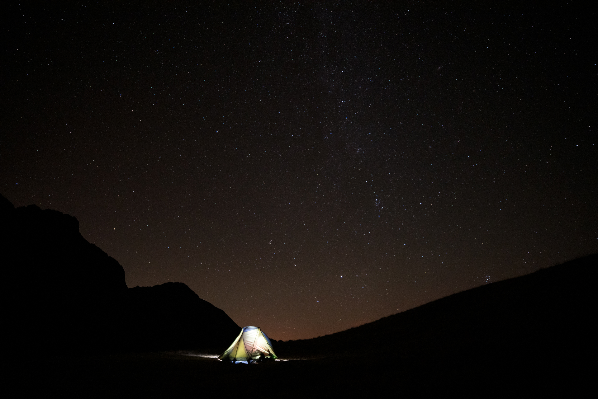 Tente de bivouac illuminée de l'intérieur, seule dans un cirque de montagne la nuit, sous un ciel étoilé avec la Voie lactée visible.