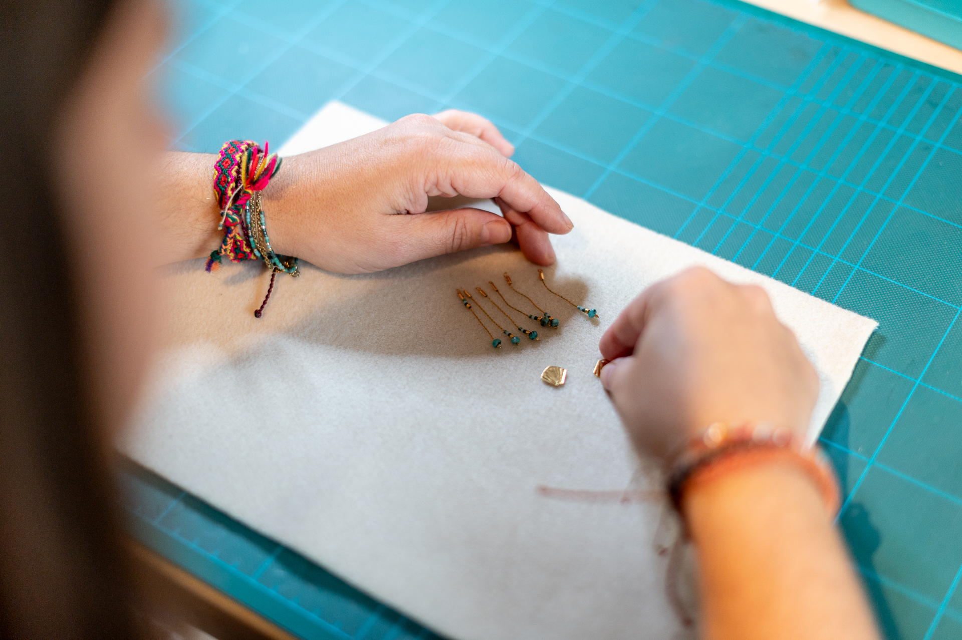 Gros plan sur les mains d'une créatrice fabriquant des bijoux, disposant des perles et pendentifs sur un tapis de découpe, pour la marque L'Otyssée.