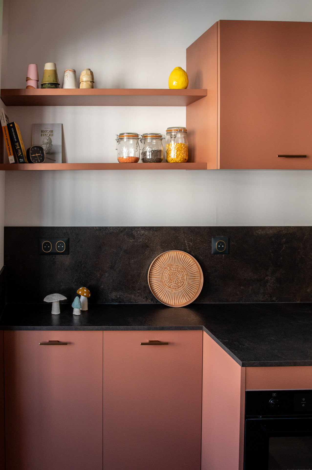 Cuisine moderne aux façades rose terracotta avec plan de travail noir, étagères assorties et objets de décoration, dans un intérieur soigné.