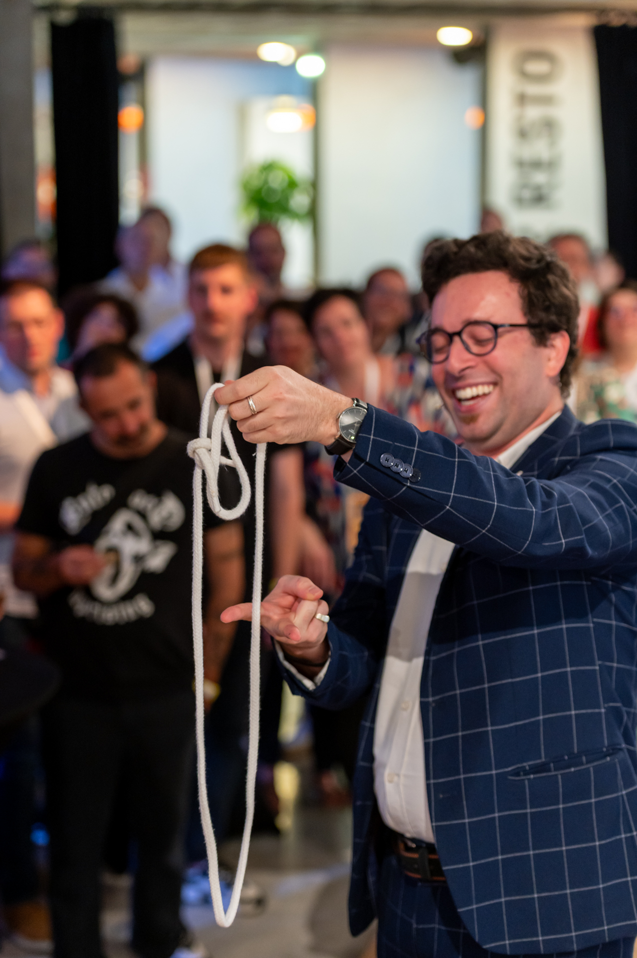 Un magicien en costume bleu réalise un tour de corde en souriant devant un public attentif, lors de la soirée des 10 ans de l'entreprise Optimistik.