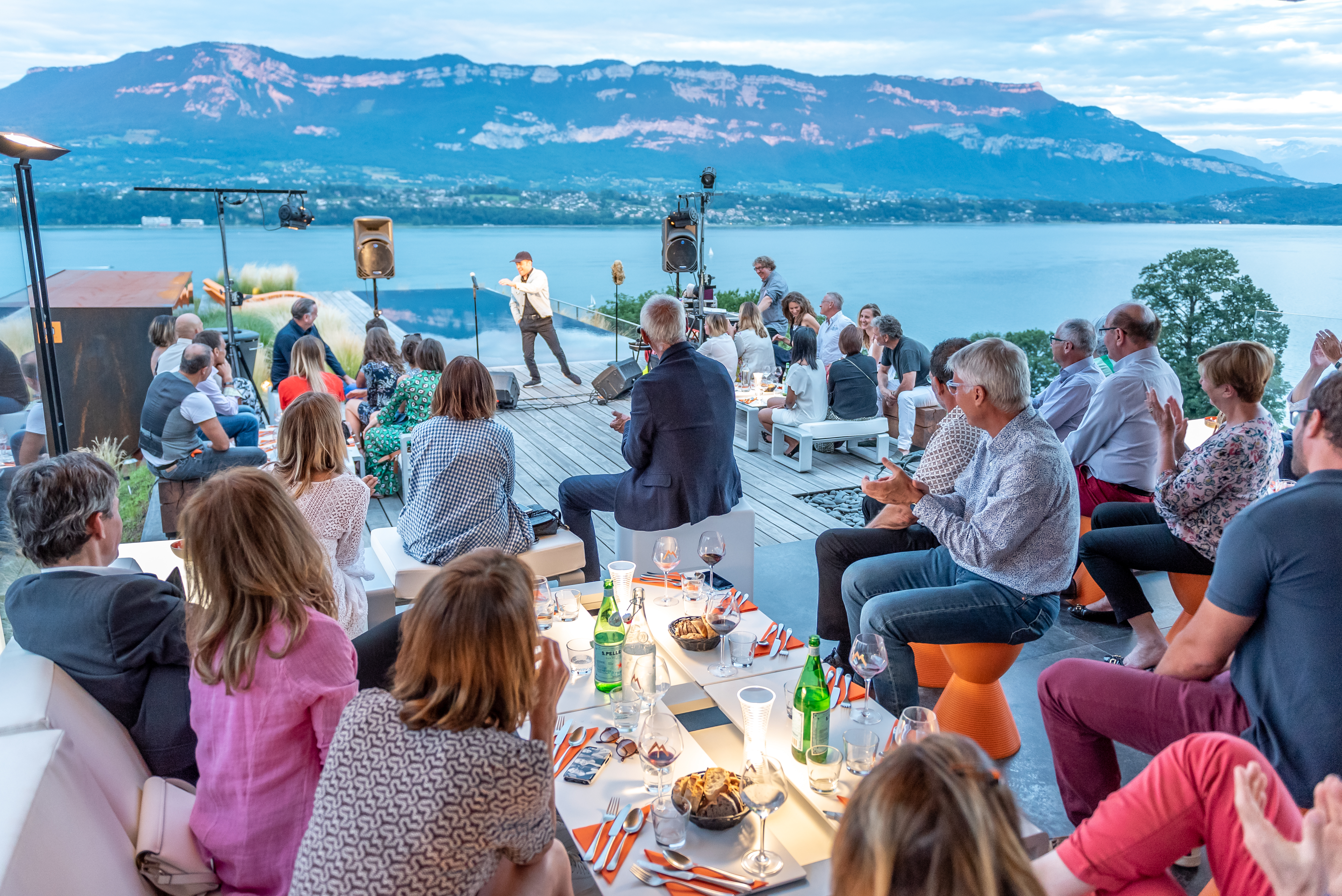 Soirée d'inauguration du JMV Resort en terrasse au bord du lac du Bourget, un public assis assiste à une prise de parole sur scène, avec la montagne éclairée par la lumière du soir en arrière-plan.