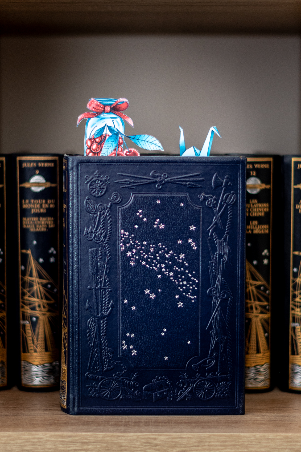 Marque-page en origami en forme de grue posé sur un livre relié bleu nuit orné d'étoiles, devant une étagère de livres anciens, création de la marque Enemesis and Art.