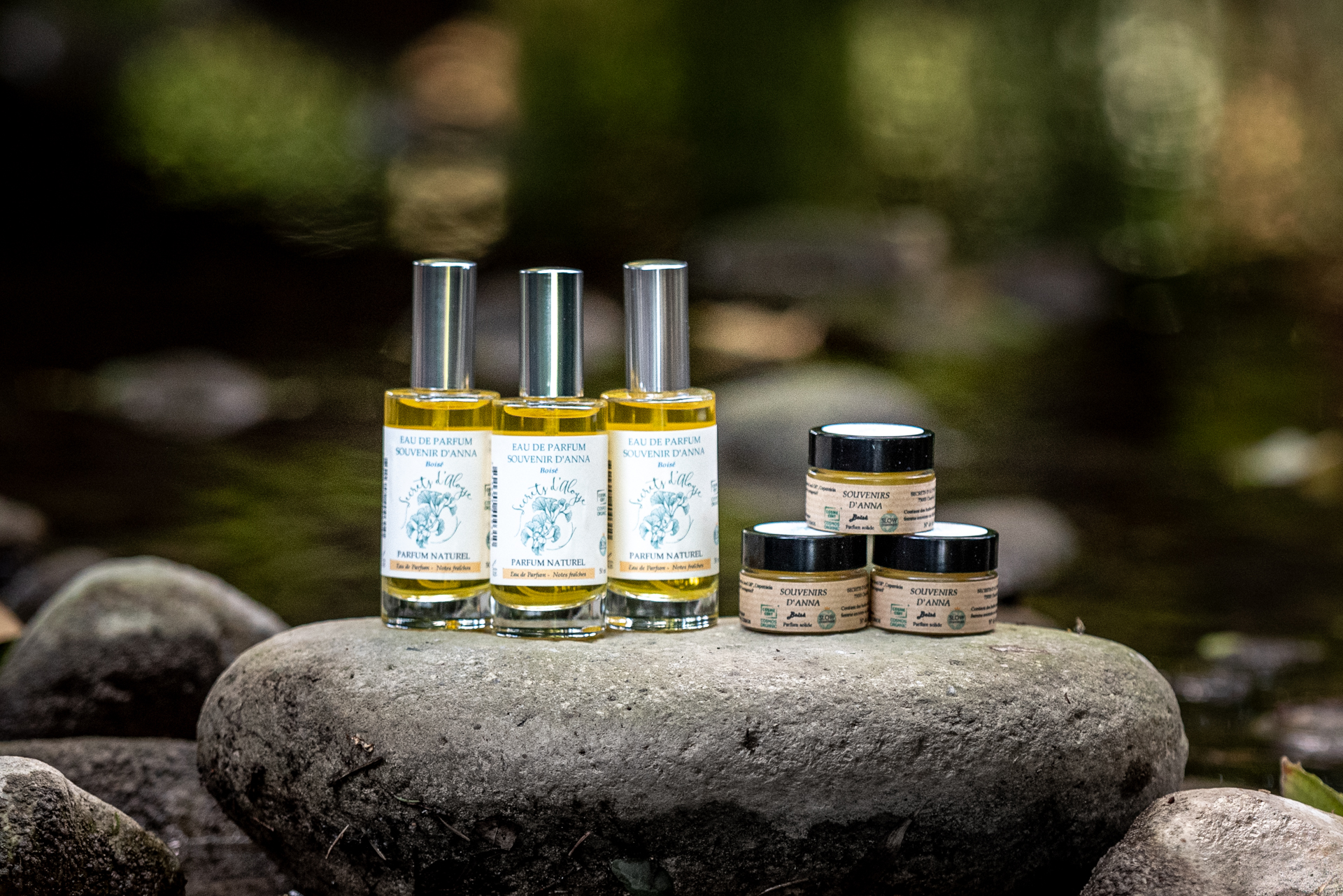 Trois flacons d'eau de parfum et deux pots de baume de la marque Secrets d'Aloyse, posés sur un rocher au bord d'un ruisseau, entourés de végétation.