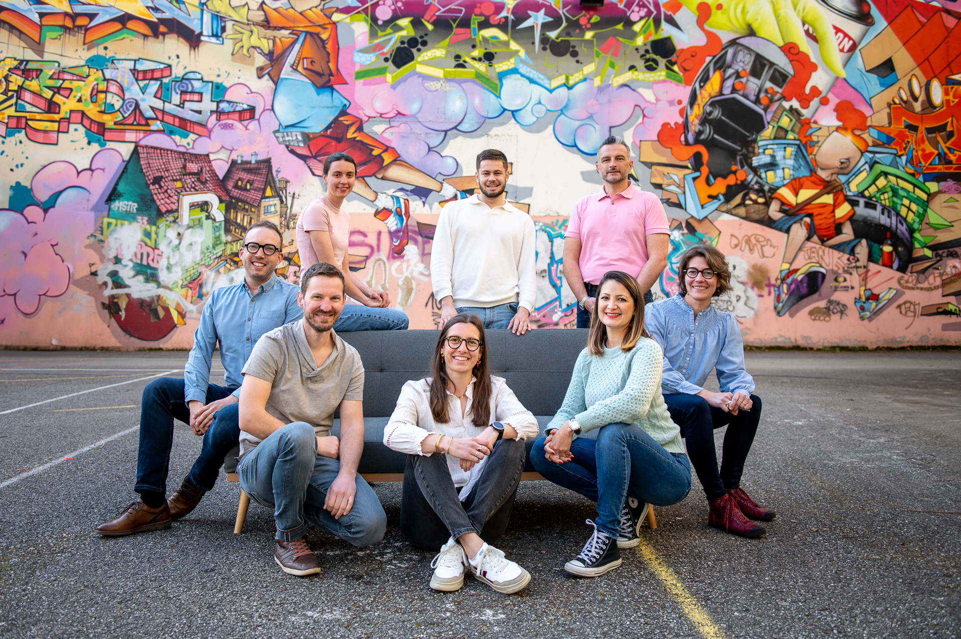 Photo de groupe de l'équipe d'Eureka Géomètres Experts, huit personnes assises et debout autour d'un canapé gris sur un parking, devant une grande fresque murale colorée en centre-ville de Chambéry.