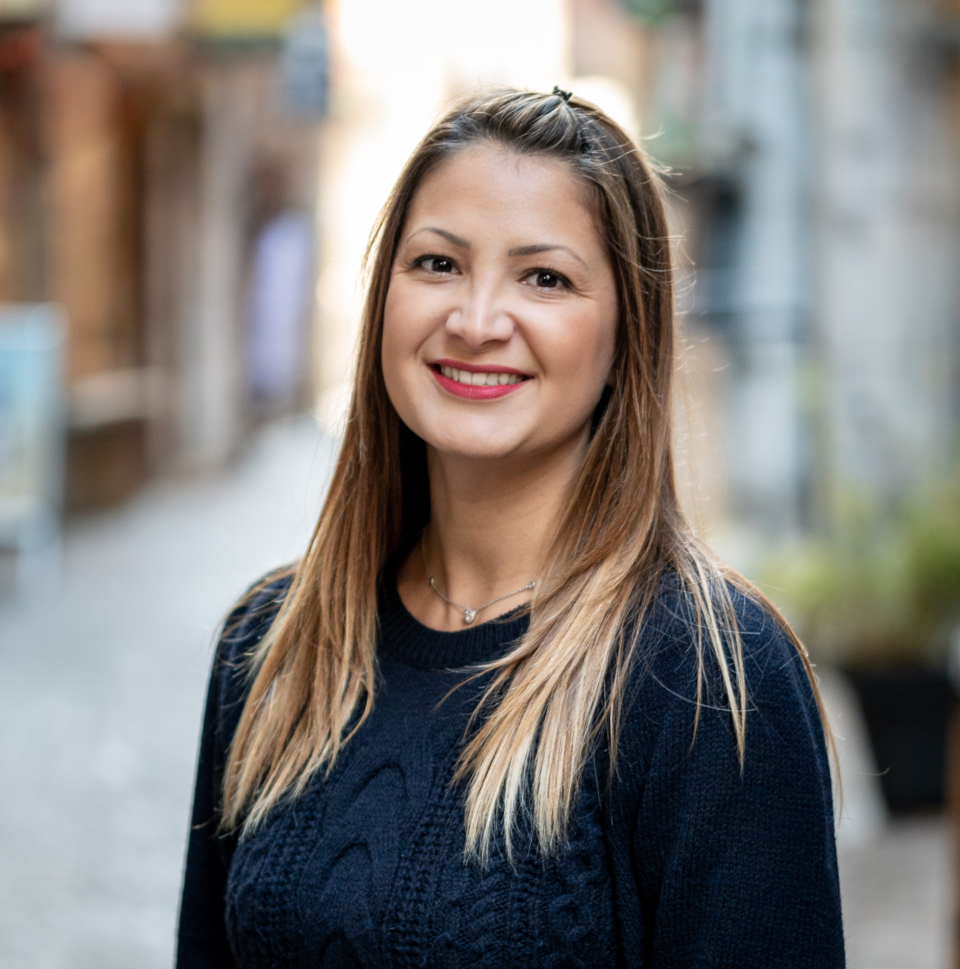 Portrait souriant d'une femme aux cheveux longs, pull noir texturé, prise en extérieur dans une ruelle pavée avec un arrière-plan flouté et lumineux.