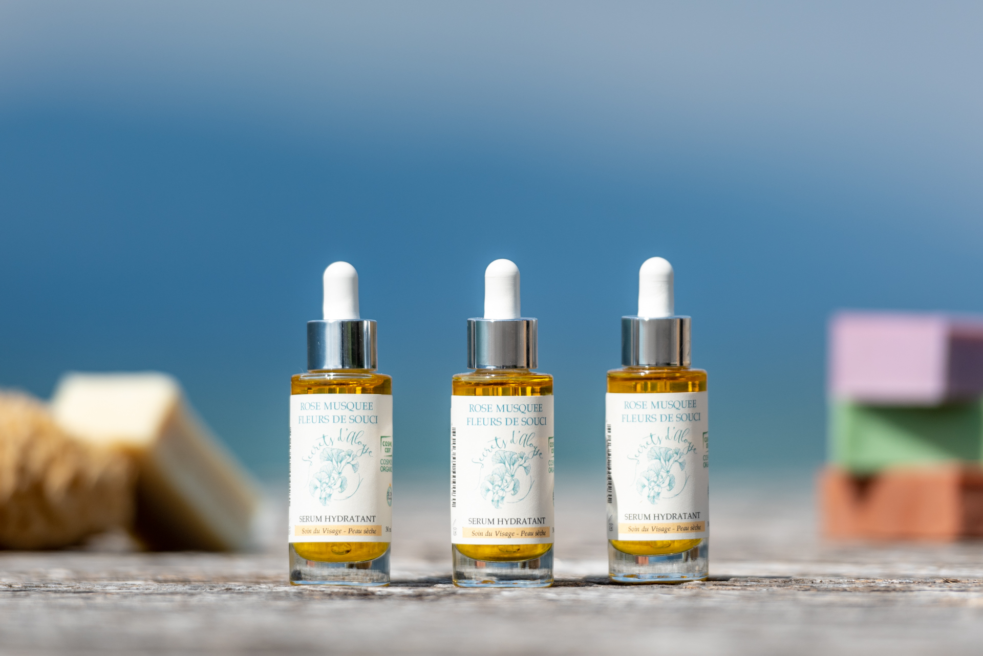 Trois flacons de sérum hydratant de la marque Secrets d'Aloyse alignés sur une surface en bois, avec des savons naturels flous en arrière-plan, fond bleu.
