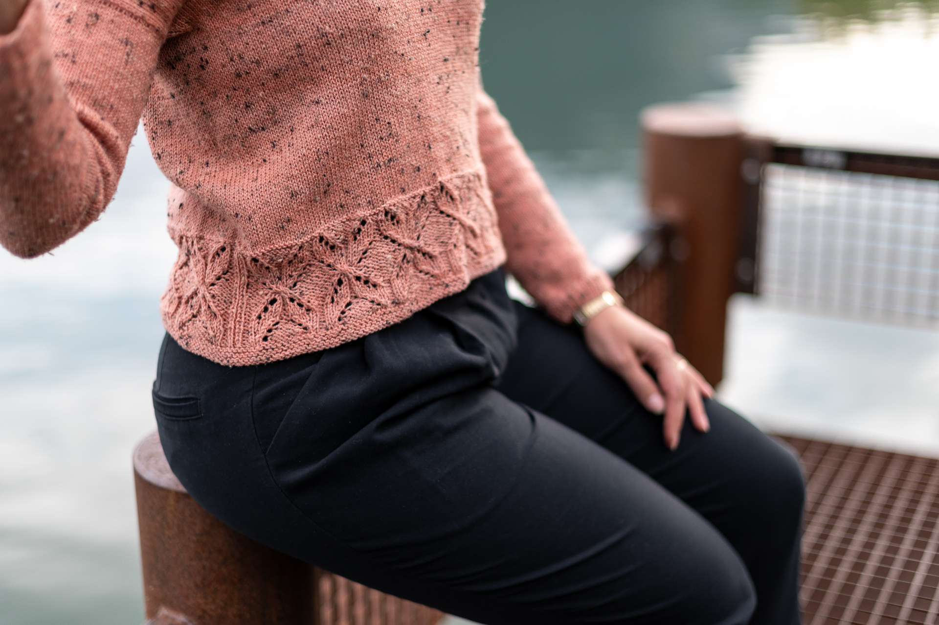 Gros plan sur un pull en tricot rose saumon avec un motif ajouré, porté par une femme assise sur un ponton en bois au bord de l'eau, création de la marque Mlle Pétronille.